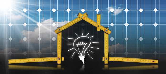 Gelber hölzerner Maßstab in der Form des ökologischen Hauses auf einem Solarpanel mit Sonnenstrahlen und einer Glühbirne.