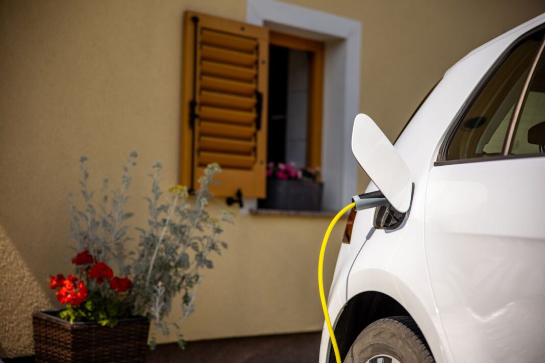 Ein weißes Auto wird über ein gelbes Kabel aufgeladen. Es befindet sich vor einem beige-gelben Gebäude mit einem blumengeschmückten Fenster und einer geöffneten, gelben Fensterklappe. Rechts sind Pflanzen.