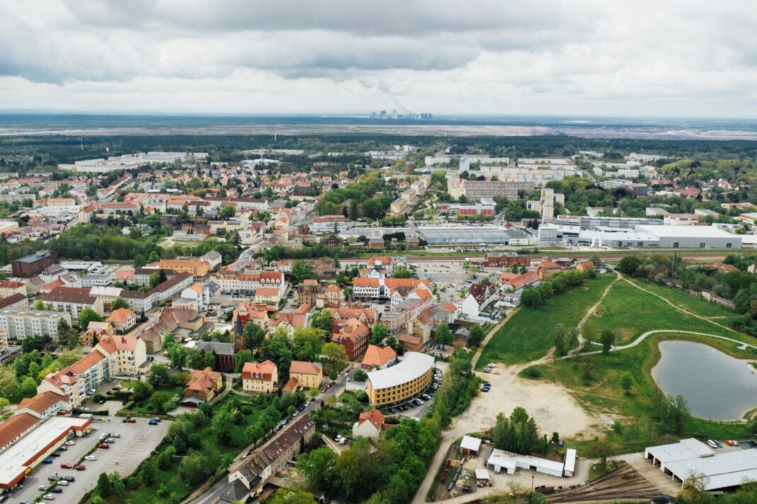 Luftaufnahme einer Stadt mit Wohnhäusern und Gewerbegebieten, flankiert von Grünflächen und einem kleinen See. Im Hintergrund zeigt sich ein bewaldeter Horizont unter wolkenverhangenem Himmel.