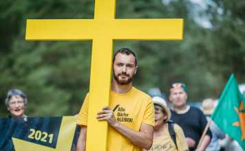 Ein junger Mann mit braunem Haar und Bart trägt ein gelbes großes Kreuz und ein gelbes T-Shirt mit der Aufschrift "Alle Dörfer leben".