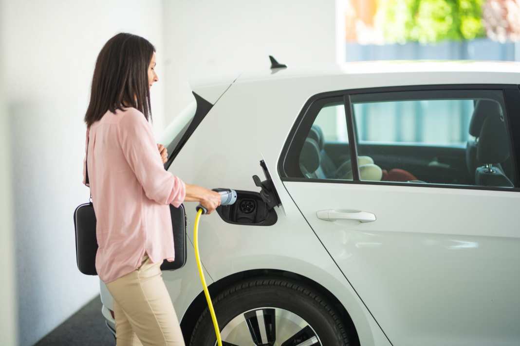 Frau lädt ein weißes Elektroauto mit einem gelben Kabel in einer hellen Garage auf.