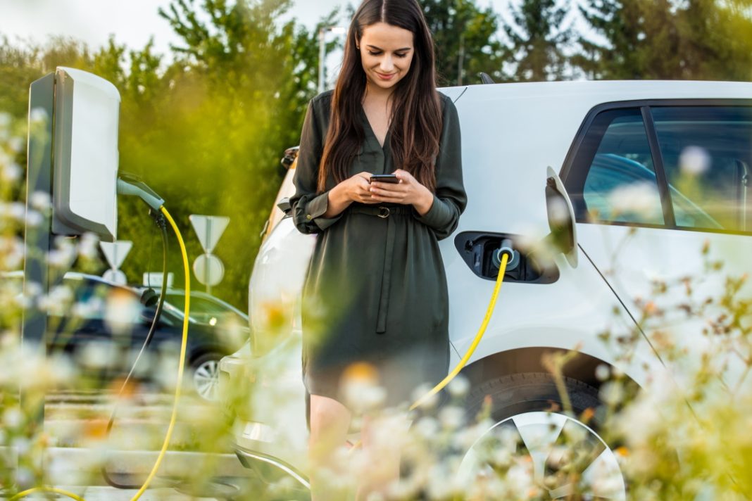 Eine Frau benutzt ihr Smartphone, während ihr Elektroauto an einer Ladesäule angeschlossen ist. Im Vordergrund sind Blumen; Bäume und Straßenschilder bilden den Hintergrund.