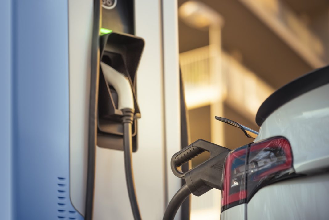 Ein Elektroauto wird an einer Ladesäule aufgeladen. Der Ladeanschluss ist an das Fahrzeug angesteckt. Die Umgebung wirkt städtisch, unscharf im Hintergrund mit Gebäudestrukturen.