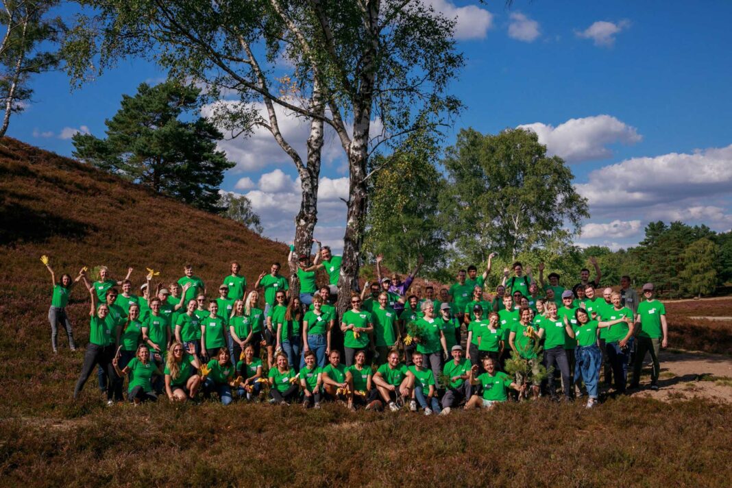 Eine große Gruppe von Menschen in grünen T-Shirts posiert lächelnd und hebt die Arme unter einem Baum im Freien. Der Himmel ist blau mit einigen Wolken.