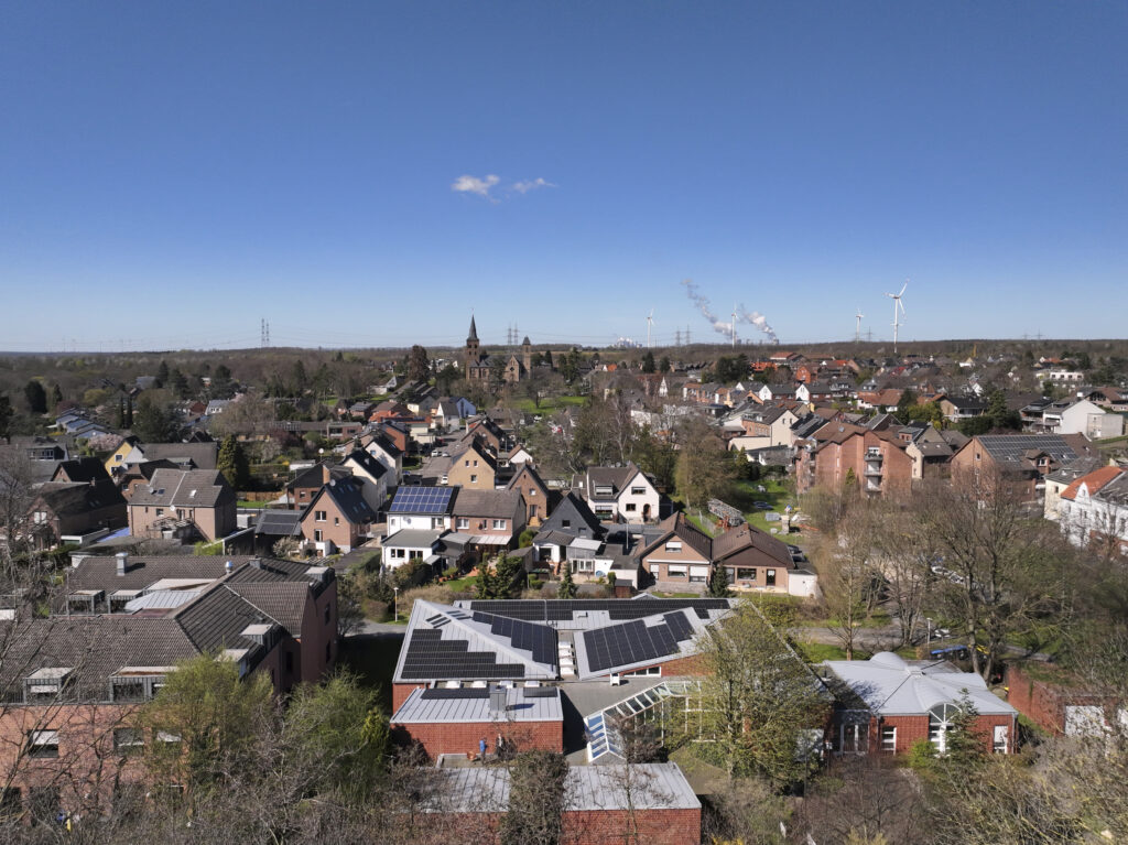 Vogelperspektive auf einen Ort, in dem auf einigen Häusern PV installiert ist. Der Himmel ist blau und wolkenlos.