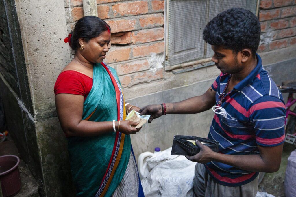 Eine Frau in einem blauen Saree zählt Geldscheine, während ein Mann in einem gestreiften Hemd sie ansieht. Sie stehen in einem schmalen, von Backsteinwänden umgebenen Durchgang.