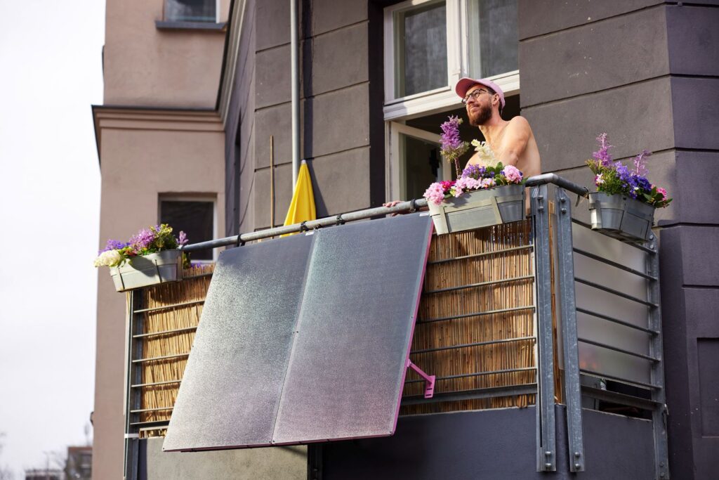 Ein Mensch auf einem Balkon gießt Blumen, umgeben von blühenden Pflanzen in Kästen. Die Balkonbrüstung ist mit Bastmatten verkleidet; eine gelbe Flagge ist sichtbar.