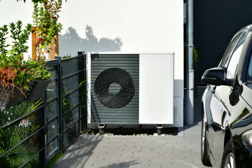 Wärmepumpe steht in der Einfahrt eines Hauses. Daneben ist ein ein Zaun mit Pflanzen und ein Teil eines schwarzen Autos zu sehen.