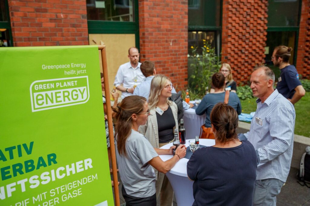 Eine Gruppe von Menschen unterhält sich an Stehtischen im Freien vor einem Gebäude. Ein grünes Plakat mit der Aufschrift „Greenpeace Energy heißt jetzt GREEN PLANET ENERGY“ ist sichtbar.