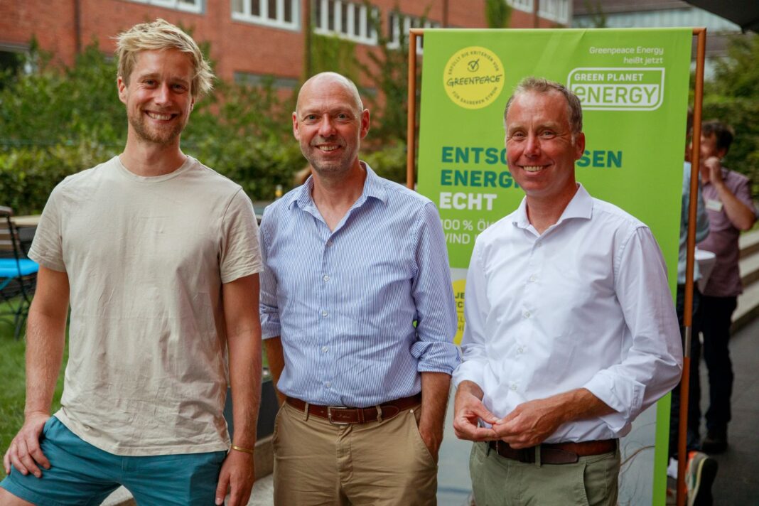 Drei Männer stehen lächelnd nebeneinander im Freien vor einem grünen Banner mit der Aufschrift „Greenpeace Energy heißt jetzt Green Planet Energy. Entschlossen. Energie. Echt.“