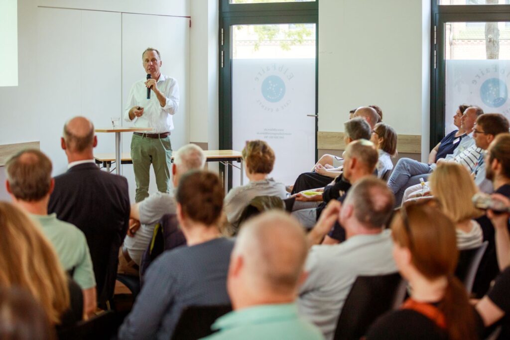 Ein Mann hält einen Vortrag vor einer sitzenden Zuhörerschaft in einem modernen Konferenzraum. Im Hintergrund sind Fenster und ein Poster mit Text sichtbar: „Eberswalde“.