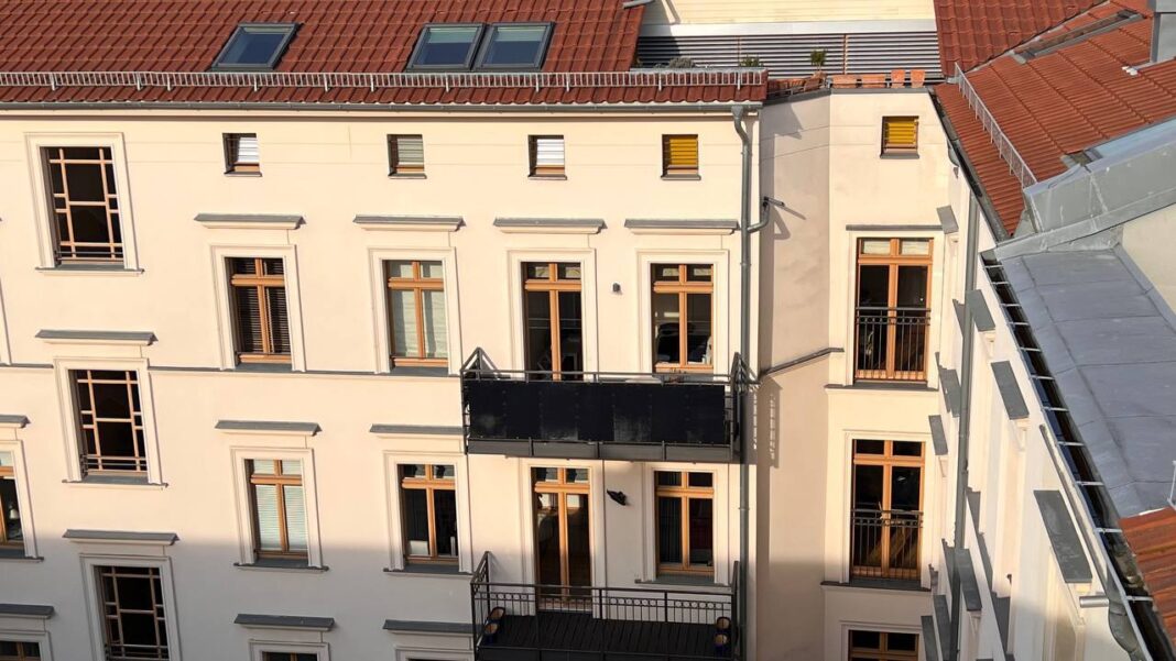 Ein mehrstöckiges Gebäude mit cremefarbener Fassade und braunen Fensterrahmen. Ein Balkon ragt heraus. Rote Ziegeldächer und Dachfenster sind sichtbar. Sonnenschein beleuchtet die Szene.