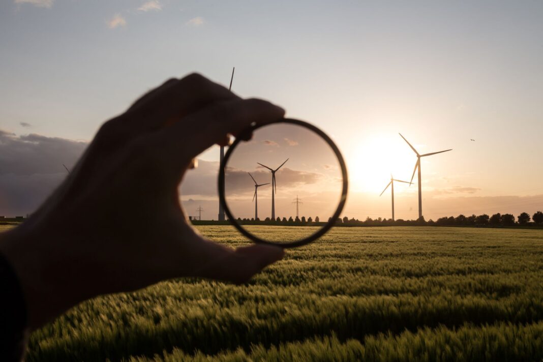 Windkraftanlagen auf einem Feld bei Sonnenuntergang. Eine im Schatten liegende Hand hält ein rundes lupenähnliches Glas vor die Windräder.