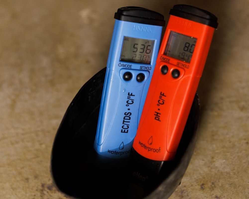 Zwei digitale Messgeräte, ein blaues und ein rotes, stehen in einem schwarzen Behälter. Auf dem Display des blauen steht „536 µS 23.7°C“, auf dem roten „8.8 pH 19.4°C“. Im Hintergrund ein heller, unscharfer Untergrund.