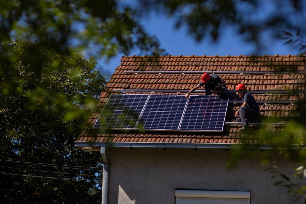 Zwei Handwerker auf dem Dach eines Einfamilienhauses beim Anbringen von PV-Modulen. Das Dach ist mit Ziegelroten Dachziegeln bedeckt. Es befinden sich bereits drei Solarmodule nebeneinander.