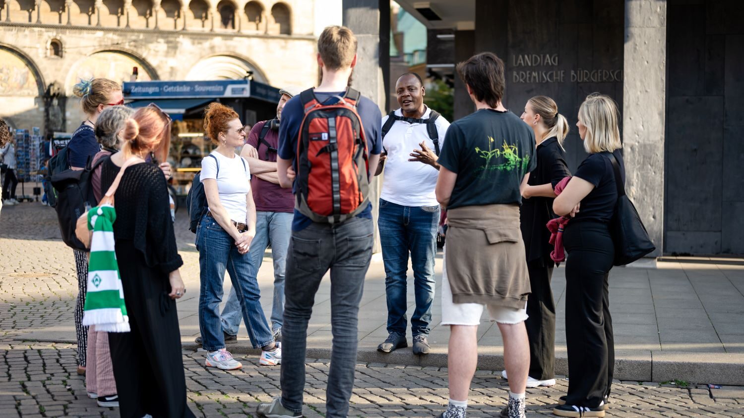 Eine Gruppe Menschen steht auf einem gepflasterten Platz im Kreis. Ein Mann in weißem T-Shirt erzählt ihnen etwas.