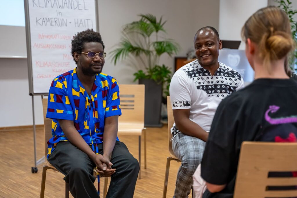 Zwei junge schwarzhaarige Männer sitzen in bunten Hemden auf Stühlen einer Person mit dunklem Shirt und blondem Dutt gegenüber. Im Hintergrund steht ein Flipchart "Klimawandel in Kamerun + Haiti. Informationen, Auswirkungen, Migration, Klimaschutz, Geschichte."