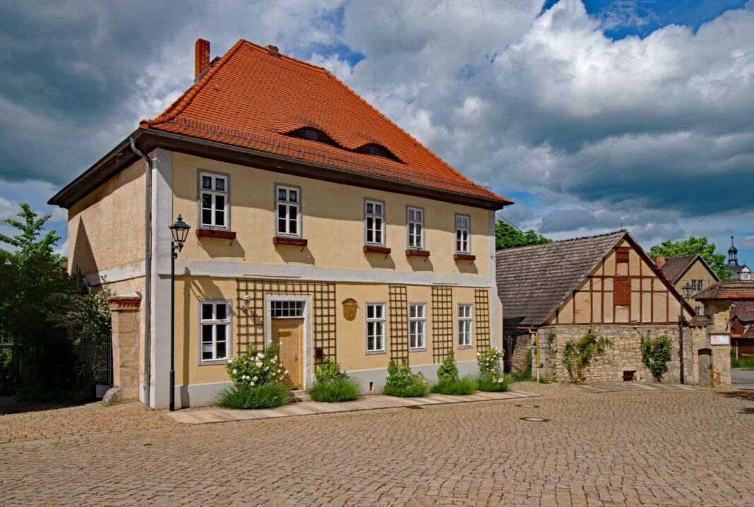 Älteres sandfarbenes Haus an einer Art Marktplatz. Das Haus hat kleine Fenster mit leeren Blumenkübeln davor. An der Hauswand sind Rankhilfen angebracht. Neben der Tür ist eine hölzerne beschriftete Tafel angebracht.