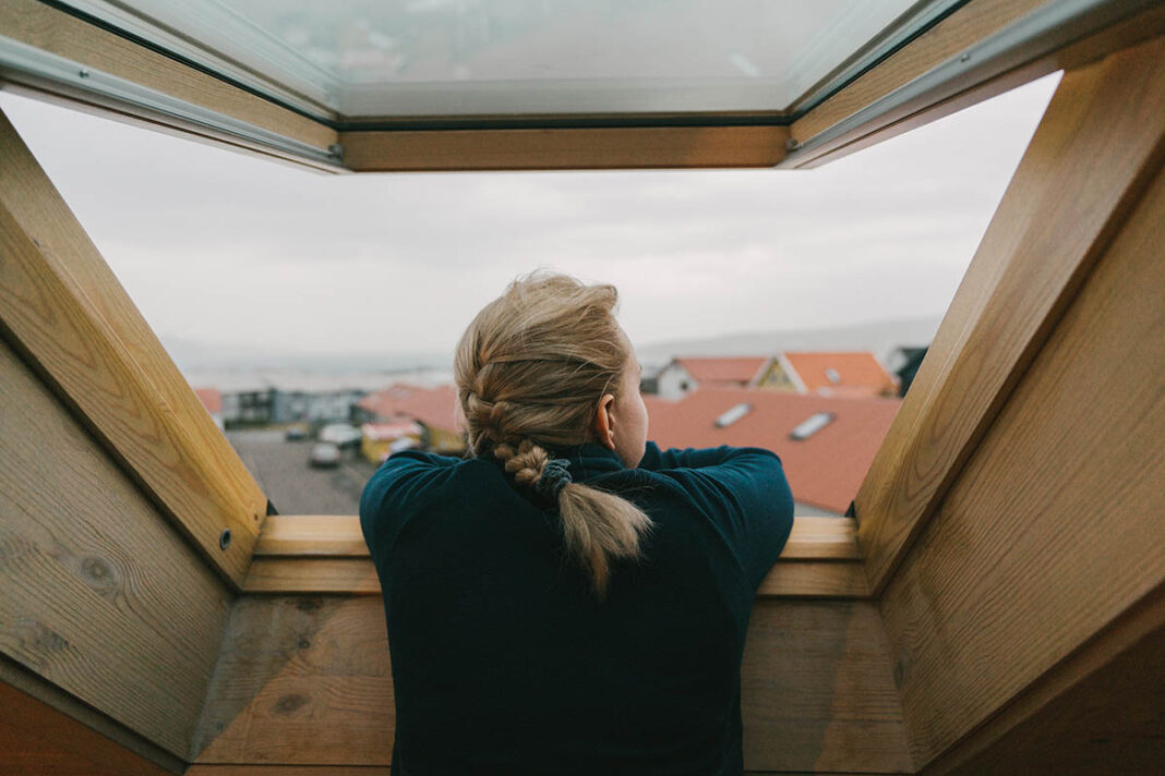 Junge Frau mit blonden, geflochtenen Haaren blickt aus einem Dachfenster auf eine Siedlung mit roten Dächern und bewölktem Himmel.