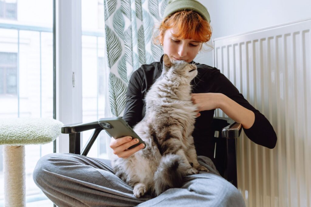 Junge Frau mit roten Haaren sitzt auf einem schwarzen Stuhl vor einer Heizung und hält ein Handy in der Hand. Auf ihrem Schoß sitzt eine Katze.