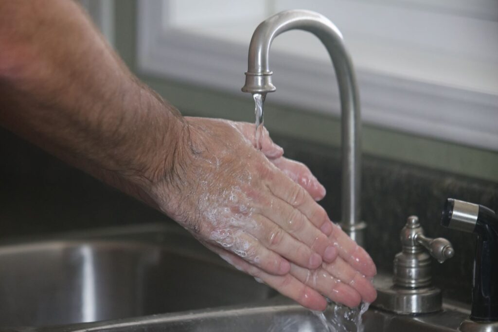 Eingeseifte Hände unter einem edelstahlfarbenen Wasserhahn. Die zugehörigen Arme sind dunkel behaart.