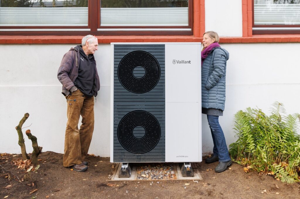 Ein Mann und eine Frau mit grauen Haaren stehen neben einer großen Wärmepumpe, die vor einem Haus steht.