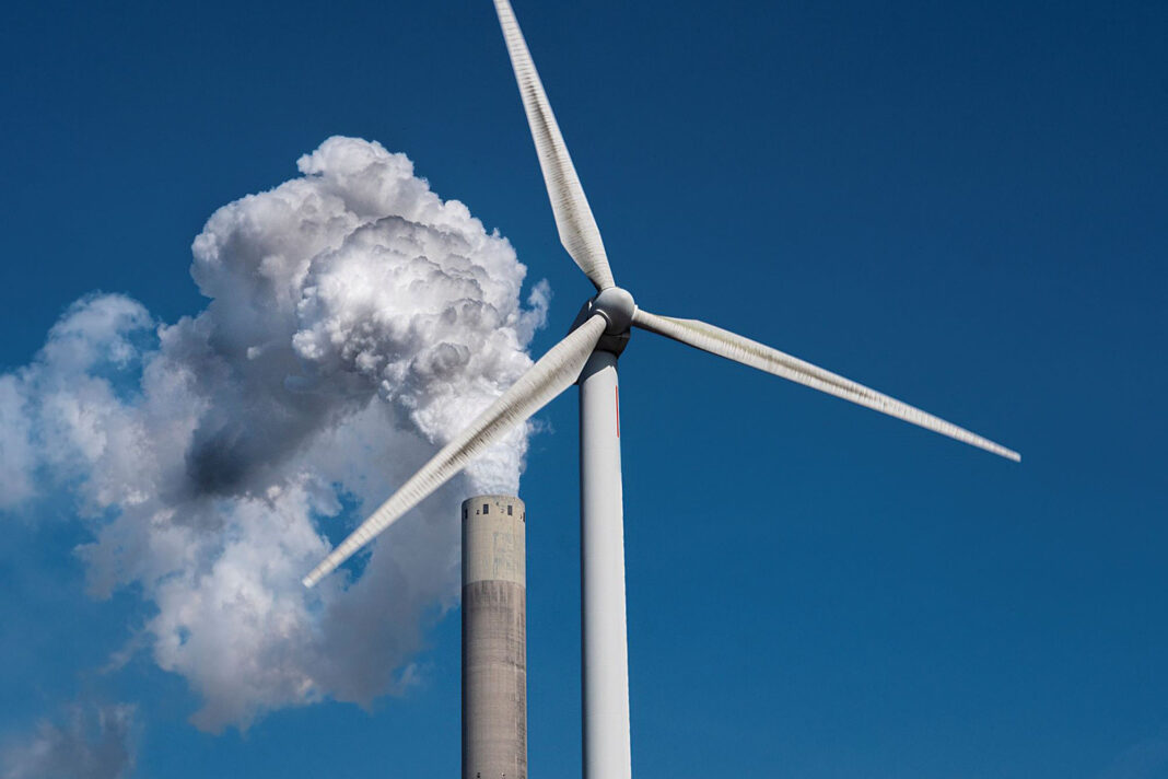 Eine Windkraftanlage und ein industrieller Schornstein mit Rauchschwaden vor einem blauen woltenfreien Himmel.