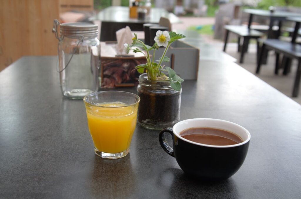 Ein Glas Orangensaft und eine Tasse Kaffee stehen auf einem Tisch in einem Café. Daneben sind ein Einmachglas mit einer Pflanze und Servietten zu sehen. Im Hintergrund Sitzgelegenheiten.