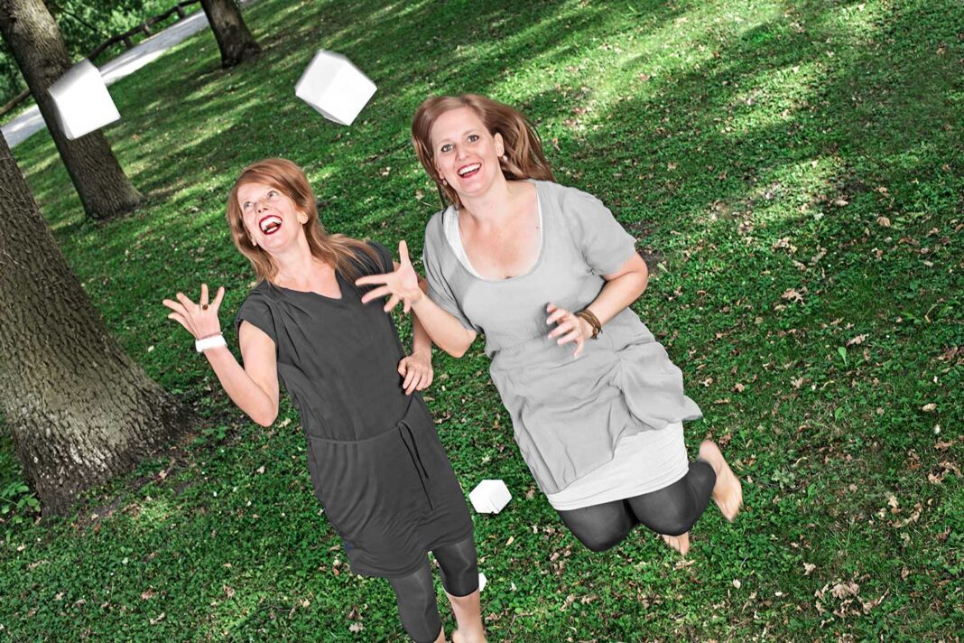 Zwei Frauen mit rotblonden Haaren, die lachend in die Luft springen und weiße Schachteln in die Höhe werfen.