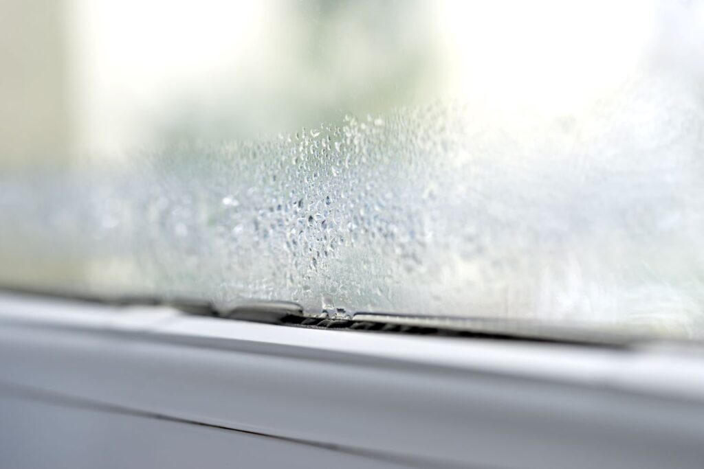 Ausschnitt eines Fensters mit weißem Fensterrahmen. Auf der Scheibe ist ein Kondenswasserstreifen zu sehen, der sich am Fensterrahmen sammelt.