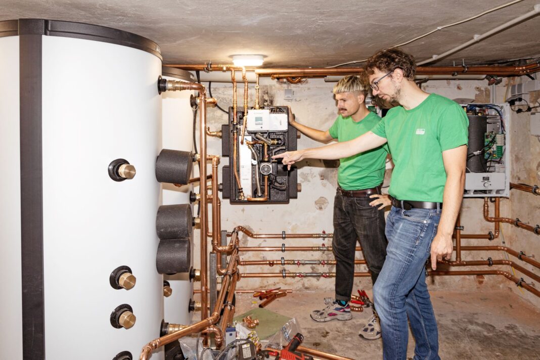 Zwei Männer stehen in einem Heizungskeller vor einer Heizungsanlage, die mit Kupferrohren im ganzen Raum verbunden ist. Die Männer tragen grüne T-Shirts mit Green Planet Energy Logo.