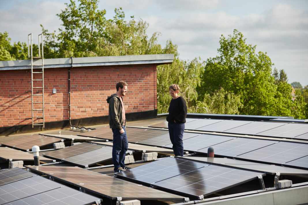 Zwei Personen in lässiger Kleidung stehen auf einem großen Flachdach, das komplett mit PV-Modulen bedeckt ist. Sie unterhalten sich lächelnd.