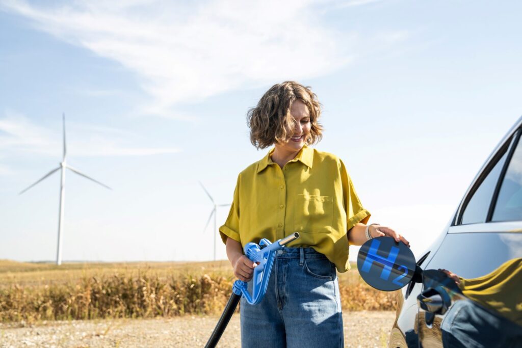 Eine junge Frau mit kurzen blonden Haaren steht an einem Weizenfeld und tankt ein Auto. Auf der Tankklappe steht "H2". Im Hintergrund sind Windräder zu sehen.