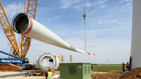 Ein Windradflügel wird von einem gelben Kran aus dem Boden gehoben. Daneben steht die Mast.