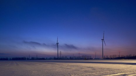 Zwei Windkraftanlagen heben sich bei Sonnenuntergang gegen den klaren Himmel ab. Der Boden ist mit Schnee bedeckt.