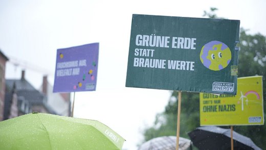 Eine Gruppe Menschen mit Regenschirmen und Demo-Schildern auf denen Slogans wie "Grüne Erde ohne braune Werte" steht.