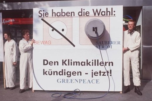 Ein altes Foto von einer Gruppe Menschen in weißen Ganzkörper-Overalls, die ein körpergroßes Schild halten auf dem "Sie haben die Wahl: Den Klimakillern kündigen - jetzt!" steht. Zusätzlich ist eine Steckdose abgebildet