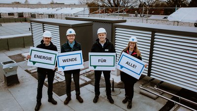 Vertreter:innen von Green Planet Energy und Fördern & Wohnen stehen auf dem Dach eines Hauses in der Wichmannstraße. Sie halten Plakate mit dem Text "ENERGIEWENDE FÜR ALLE!"