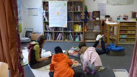 Kinder sitzen in einem bunten Klassenzimmer auf dem Boden und beschäftigen sich mit Zetteln, die vor ihnen liegen.