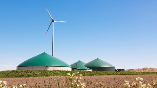 Eine Biogasanlage hinter einem Ackerfeld. Dahinter steht eine Windkraftanlage.