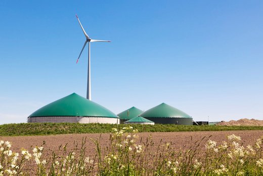 Eine Biogasanlage hinter einem Ackerfeld. Dahinter steht eine Windkraftanlage.