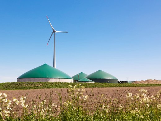 Eine Biogasanlage hinter einem Ackerfeld. Dahinter steht eine Windkraftanlage.