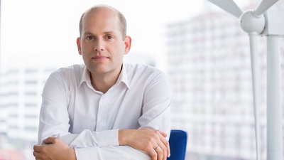 Ein Mann in einem weißen Hemd sitzt an einem Tisch, stützt seine Arme darauf und schaut in die Kamera. Im Hintergrund ist ein Modell einer Windturbine vor unscharfen Gebäuden sichtbar.