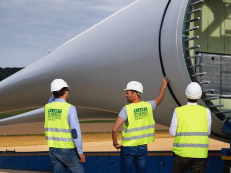 Drei Arbeiter in Warnwesten stehen vor einem Flügel einer Windanlage.