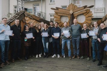 Sechzehn Personen stehen in einem Halbkreis und blicken in die Kamera, manche halten ein DINA4 Blatt vor sich