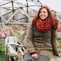 Eine Frau sitzt lächelnd in einem braunen Mantel und rotem Schal auf einer Holzbank im Freien. Im Hintergrund befinden sich eine metallische Kuppelstruktur und unscharfe Gebäude.