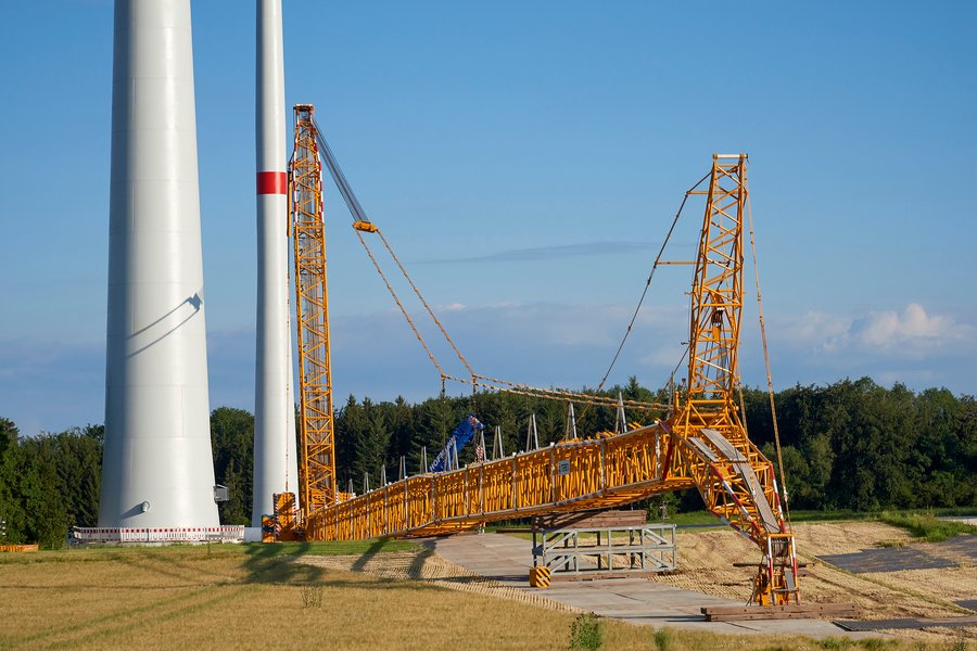 Ein großer gelber Kran montiert einen Windkraftturm. Im Hintergrund stehen Bäume und unter einem blauen, leicht bewölkten Himmel. Der Kran befindet sich auf einer weiten, grasbewachsenen Fläche.