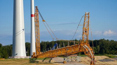 Ein großer gelber Kran montiert einen Windkraftturm. Im Hintergrund stehen Bäume und unter einem blauen, leicht bewölkten Himmel. Der Kran befindet sich auf einer weiten, grasbewachsenen Fläche.