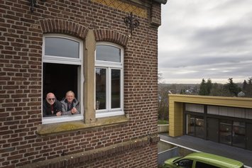 Drohnenaufnahme von einem Gebäude. Zwei Männer sind an ein offenes Fenster über einen Fensterrahmen gelehnt 