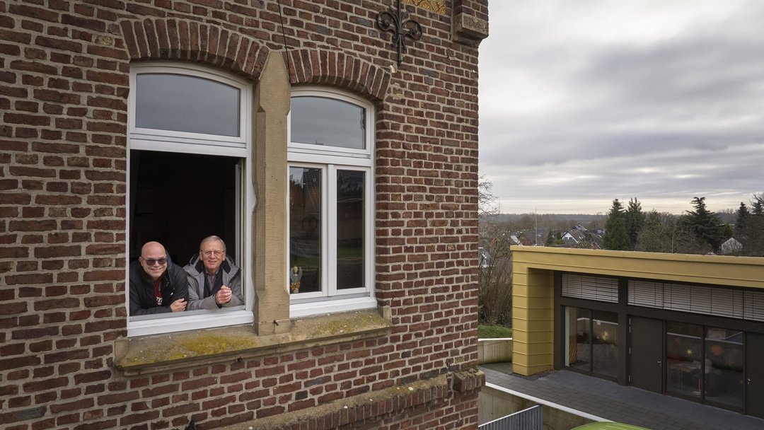 Drohnenaufnahme von einem Gebäude. Zwei Männer sind an ein offenes Fenster über einen Fensterrahmen gelehnt